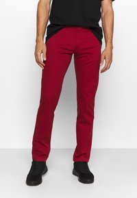 Pantaloni di cotone rossi con vestibilità slim, dotati di tasche laterali e passanti per la cintura, abbinati a sneakers nere. Tessuto liscio e colore uniforme.