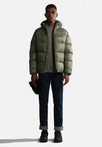 Giacca a vento verde con cappuccio, chiusura con zip e design trapuntato. Indossata sopra una felpa verde coordinata, abbinata a jeans blu scuro e scarpe nere.