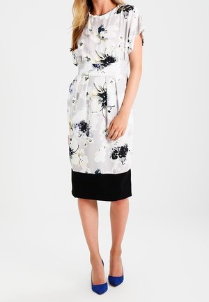 Mujer con vestido blanco floral hasta la rodilla con bajo negro y tacones altos azules, de pie frente a un fondo claro y liso.