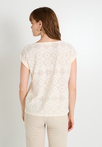 Blusa de manga corta color crema con un patrón de encaje floral y puños enrollados, con un corte holgado y un diseño de espalda abierta para mayor transpirabilidad.