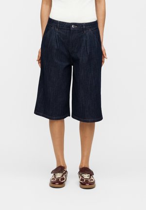 Personne portant un short en jean bleu foncé jusqu’aux genoux, un haut blanc, et des baskets marron avec des lacets crème, debout sur un fond uni.