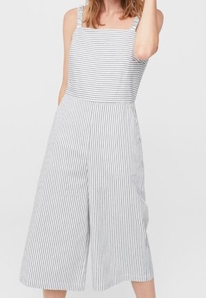 Femme portant une combinaison sans manches à jambes larges avec des rayures verticales et horizontales grises et blanches, debout devant un fond uni.