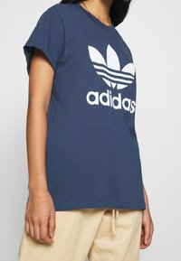 Tmavě modré bavlněné tričko s krátkými rukávy a kulatým výstřihem. Na přední straně se nachází bílé logo Adidas a design tří listů.