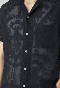 Camicetta nera a maniche corte in rete con motivi floreali, davanti con bottoni, taschino sul petto, indossata sopra una maglietta bianca e una sottile collana in argento.