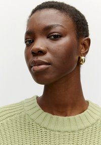 Strickpullover in Hellgrün mit strukturiertem Muster und ribbeltem Ausschnitt; das Modell trägt goldene kreisförmige Ohrringe und hat kurzes, natürliches Haar.