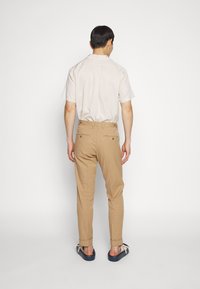 Beige kortärmad skjorta, beige byxor och marinblå sandaler. Byxorna har två bakfickor med knappstängning. Enkel design och avslappnad passform.