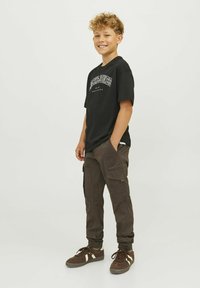 Čierne tričko s bielou grafikou, v kombinácii s hnedými cargo nohavicami s bočnými vreckami a rebrovanými manžetami; hnedé tenisky s bielymi akcentmi.