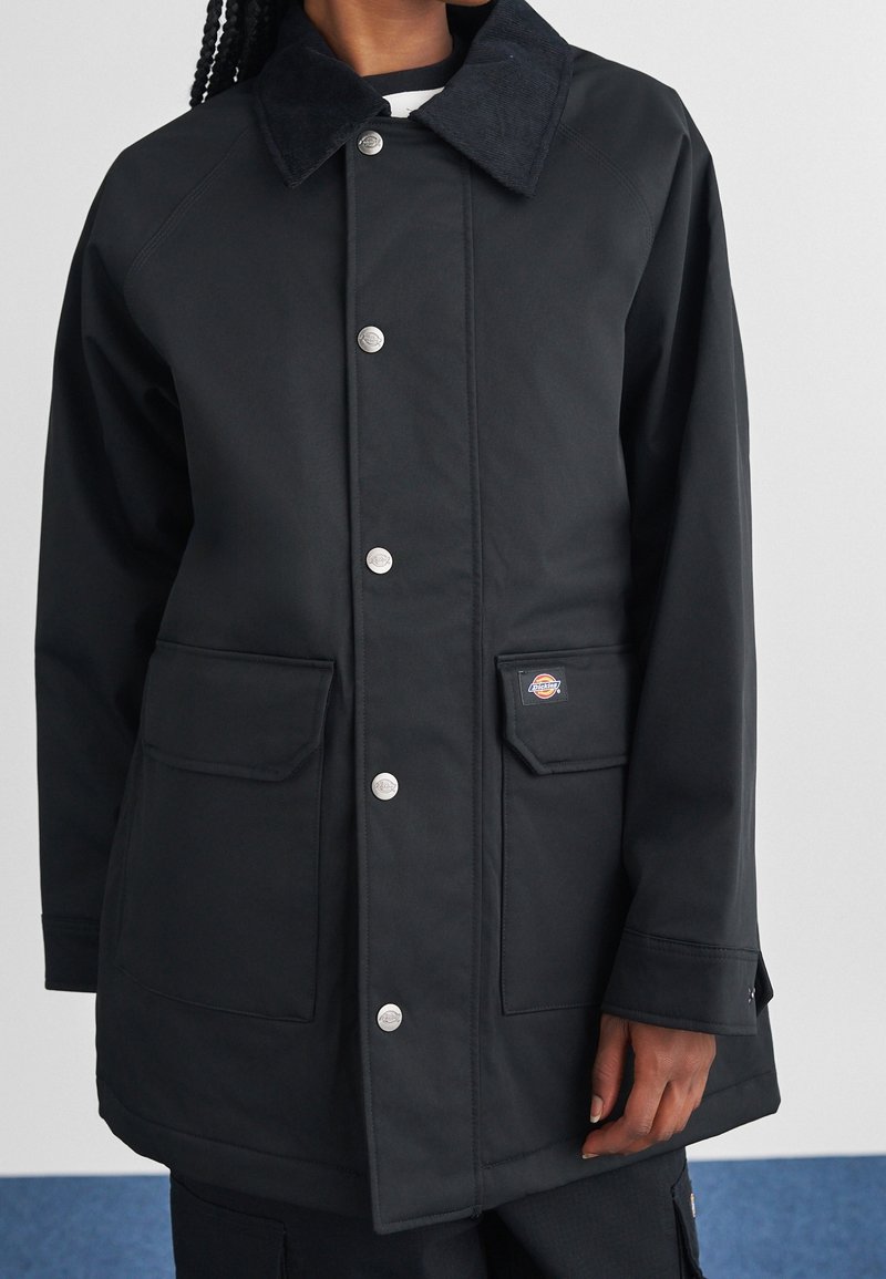 Personne portant un manteau noir à boutons avec de grandes poches et un petit patch logo sur une poche, debout devant un fond uni.