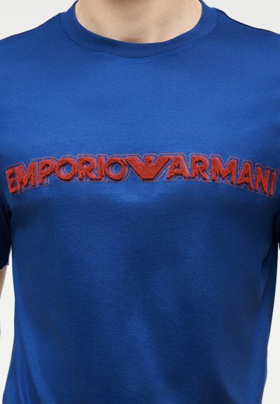 Μπλε μπλουζάκι με στρογγυλή λαιμόκοψη, με κόκκινη, ανάγλυφη κεντημένη επιγραφή "Emporio Armani" και λογότυπο αετού στο ύψος του στήθους.