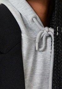 Grijze hoodie met een gestructureerde afwerking, voorzien van een trekkoord met een witte plastic stopper, gedeeltelijk bedekt door een zwarte buitenlaag.