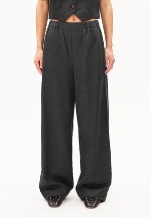 Vrouw in donkergrijs wijde broek met elastische tailleband, bijpassend cropped vest en zwarte gestoorde schoenen tegen een witte achtergrond.
