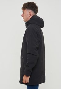 Giacca nera con cappuccio, caratterizzata da una texture liscia e opaca. Presenta tasche laterali con zip e una vestibilità comoda, che arriva fino a metà coscia.