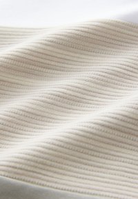 Gaiši bēšas krāsas teksturēts audums ar smalku ribu rakstu, rādot maigas locījumu līnijas un gludu virsmu, kas pāriet baltā malā.