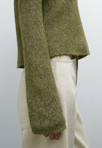 Person wearing olive green knitted sweater with long sleeves and light beige pants, shown from the side below the waist.
