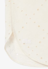 Tissu crème avec de petits trous ajourés et motifs d’alligator brodés, comportant un logo alligator cousu près de l’ourlet incurvé.