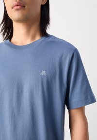 Světle modré bavlněné tričko s kulatým výstřihem a krátkými rukávy. Výrazná malá bílá výšivka "MOP" na hrudi. Hladká textura.