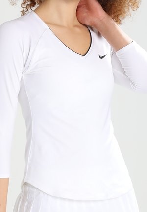 Femme portant un haut de sport blanc à manches longues avec col en V et logo Nike noir, touchant son cou, cadré aux épaules et à la taille.