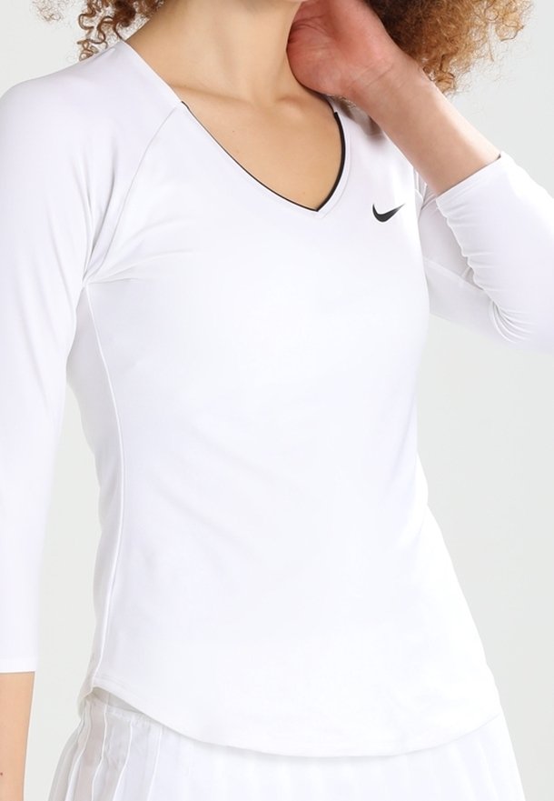Femme portant un haut de sport blanc à manches longues avec col en V et logo Nike noir, touchant son cou, cadré aux épaules et à la taille.