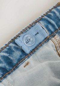 Tissu en denim bleu avec un léger effet délavé, doté d'une étiquette en tissu et d'un bouton transparent. Accents de couture en brun clair sur les bords.