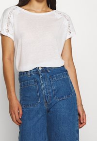 Bílý krátký rukáv top s krajkovými detaily na ramenech, v kombinaci s modrými džíny s vysokým pasem, které mají velké kapsy a klasický střih.