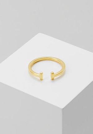 Bague ouverte en or avec des extrémités plates en forme de barre verticale reposant sur un piédestal cubique blanc sur un fond gris uni.