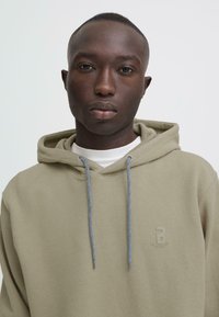 Hoody in olijfgroen, gestructureerde stof, met een logo op de linkerborst en contrasterende blauwe trekkoorden. Draagt over een wit shirt.