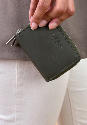 Green leather wallet with a zip closure, featuring a subtle embossed logo and smooth texture, held in a hand against light-colored fabric.