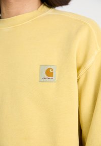 Sweatshirt jaune avec une étiquette logo ronde aux couleurs orange et blanche. Le tissu semble doux avec des détails de couture visibles.