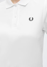Polo blanc avec col et deux boutons, avec un petit logo noir de couronne de laurier sur la poitrine gauche.