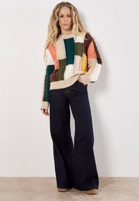 Sweter dzianinowy z wielokolorowym wzorem patchworkowym i teksturą warkocza; w połączeniu z szerokimi ciemnymi dżinsami i beżowym obuwiem.