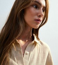 Beige skjorta med krage och strukturerad yta samt två knappar, bärs med ett delikat silverhalsband mot en ljus bakgrund.