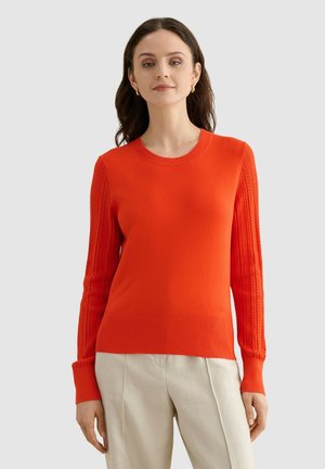 Mujer con cabello oscuro que lleva un suéter naranja de manga larga con mangas texturizadas y pantalones beige, de pie frente a un fondo liso.