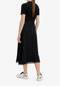 Schwarzes Rippenkleid mit einem gestuften Spitzenabschluss, kombiniert mit weißen Sneakern mit schwarzen Texteinakzenten. Glatte Textur, knielange.