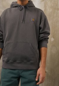 Persona che indossa una felpa Carhartt grigio scuro con tasca frontale e cordini, abbinata a pantaloni teal, in piedi contro un muro texture.