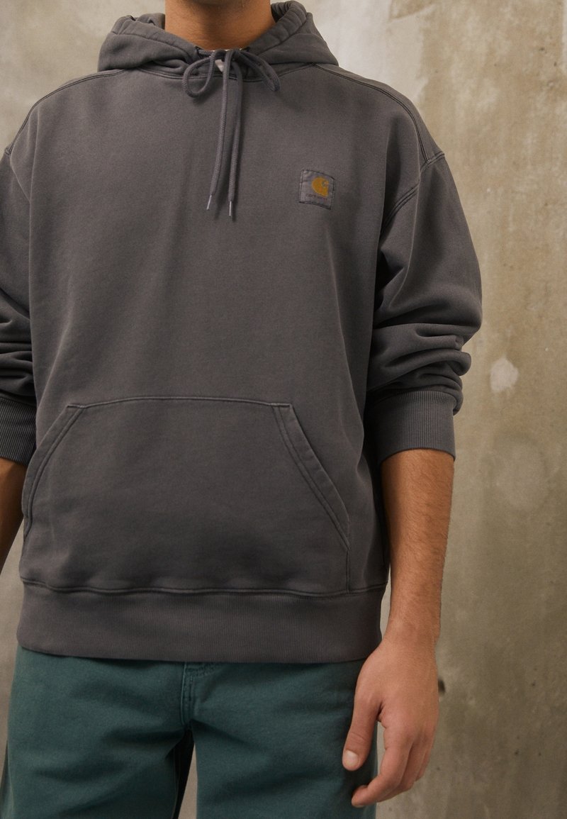 Persona che indossa una felpa Carhartt grigio scuro con tasca frontale e cordini, abbinata a pantaloni teal, in piedi contro un muro texture.