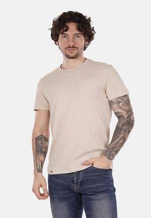 T-shirt beige a maniche corte realizzato in tessuto morbido, con una tasca sul petto testurizzata e un collo a giro. Abbinato a jeans blu.