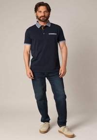 Polo shirt marine avec un col et des poignets bleu clair, patte de boutonnage à trois boutons et une poche poitrine. Associé à un jean foncé et des baskets claires.