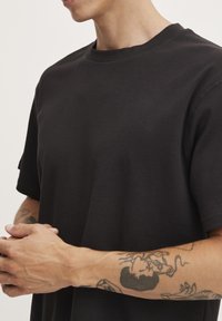 Camiseta negra con cuello redondo clásico, mangas cortas y una textura suave, llevada por una persona con tatuajes visibles en los brazos.