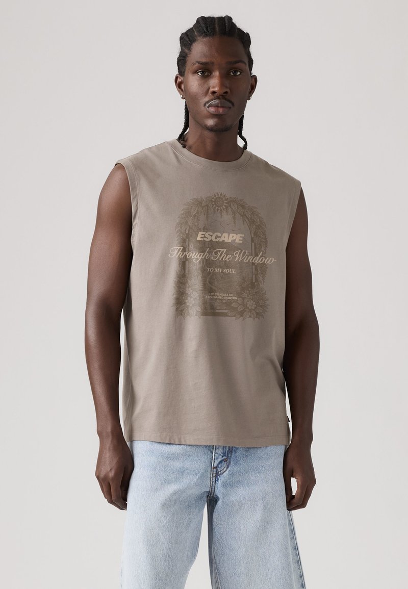 Mann mit geflochtenem Haar, der ein ärmelloses beiges Shirt mit floralem Fenster-Design und dem Text „Escape Through The Window“ trägt, kombiniert mit hellblauen Jeans.