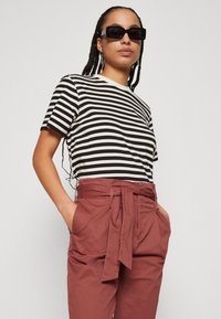 Schwarz-weißer gestreifter T-Shirt mit kurzen Ärmeln, kombiniert mit hoch geschnittenen, beige Hosen mit Bindegürtel. Das Model trägt schwarze Sonnenbrillen.