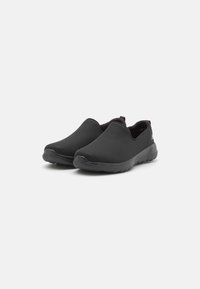 Zapatos negros tipo slip-on con una parte superior de malla texturizada, punta redonda y una suela acolchada para mayor comodidad. Diseño minimalista sin herrajes visibles.