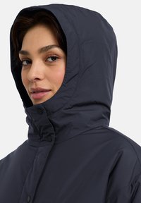 Giacca puffa blu navale con texture trapuntata, colletto alto e grande cappuccio. Presenta bottoni a pressione neri sulla parte anteriore per la chiusura.