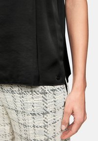 Schwarzes, glattes Oberteil mit seitlichen Schlitzen; kombiniert mit strukturierten, gemusterten beige-schwarzen Hosen, die ein gewebtes Design zeigen.