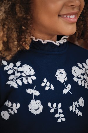 Navy sweater with white floral embroidery and scalloped neckline. Features leaf and flower patterns on the front with ribbed fabric texture.