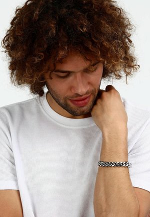 Bracciale a catena color argento indossato al polso, con maglie intrecciate e finitura lucida. Lo sfondo è bianco luminoso.