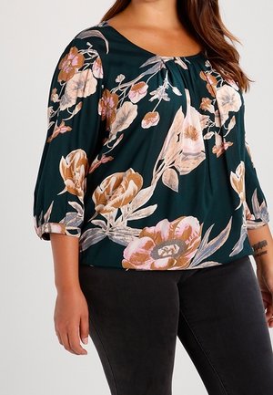 Kobieta w ciemnozielonej bluzce z różowo-beżowym kwiatowym wzorem i czarnych spodniach, stojąca z lewym ramieniem swobodnie opuszczonym wzdłuż ciała.
