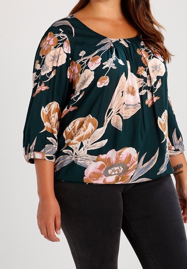 Femme portant un chemisier vert foncé à imprimé floral rose et beige et un pantalon noir, debout avec son bras gauche détendu le long du corps.