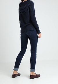 Marineblå stickad hoodie med dragsko, matchad med mörkblå jeans som är uppvikta vid anklarna och vinröda slip-on skor. Enkel och figurnära design.