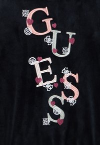 Gucci logo izvezen na crnoj tkanini s ružičastim i bijelim slovima ukrašenim malim bordo srcima i bijelim kružnim motivima.