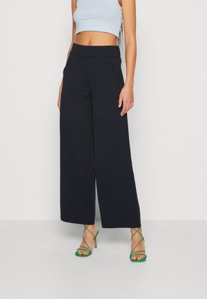 Woman wearing black wide-leg pants, a light blue cropped top, and green strappy high-heeled sandals, standing against a plain background.